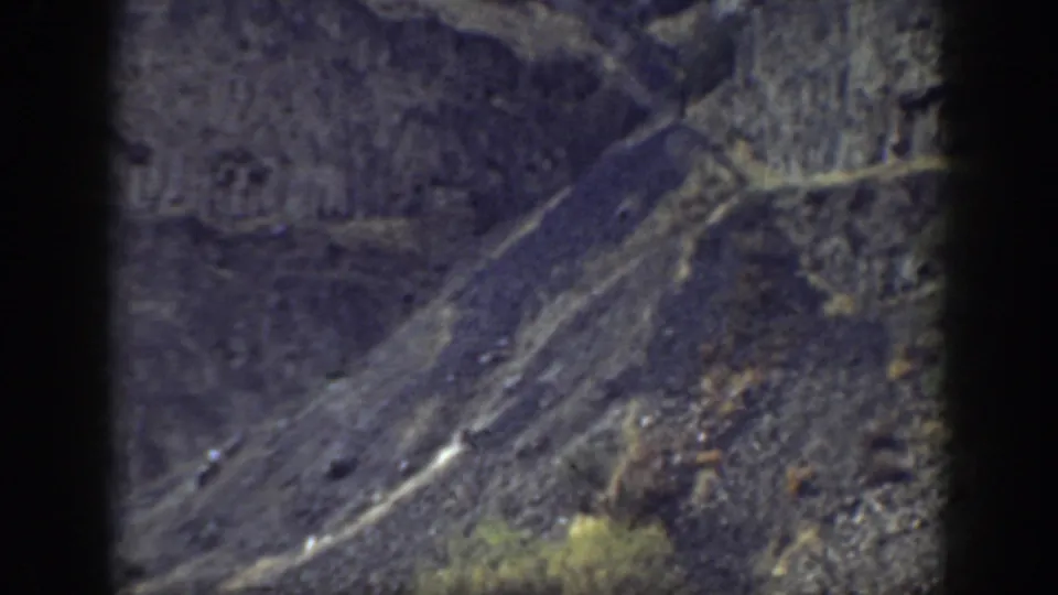 1937 California home-movie frame of a rugged hillside, with layered rock, scree, pale diagonal traces, and sparse vegetation.
