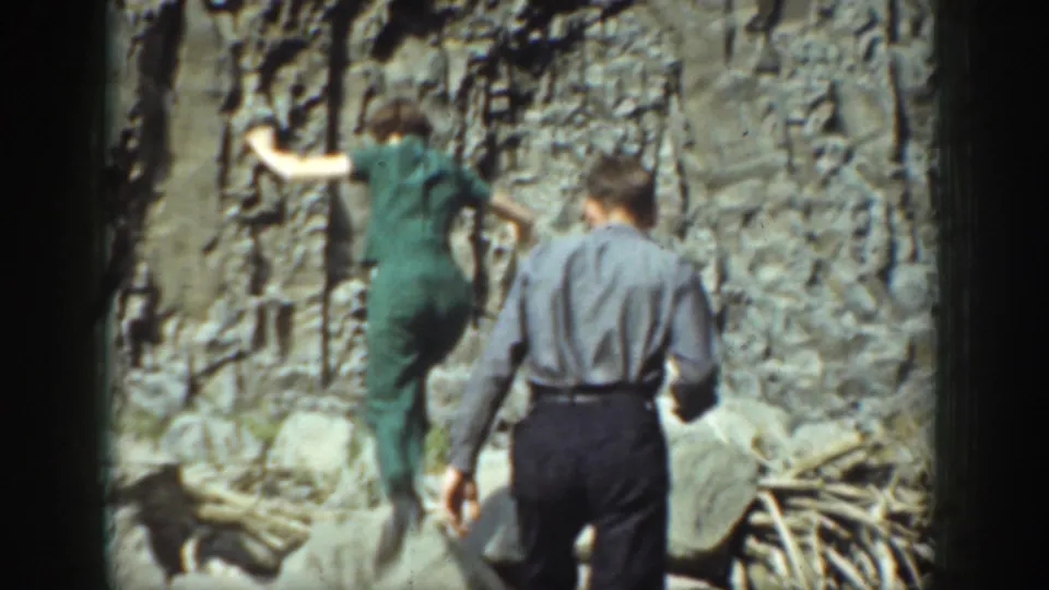 1937 California home-movie frame showing an adult woman climbing a rock formation as an adult man follows behind.