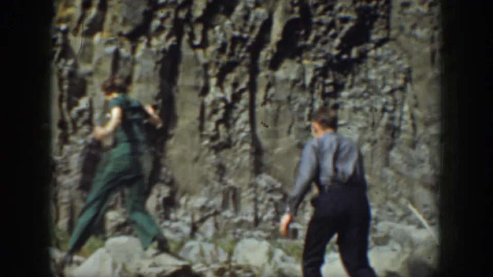 1937 California 8mm frame of a woman in green and a man in dark trousers crossing rocky terrain.