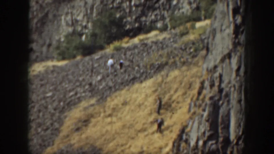 1937 California home-movie frame showing distant adult figures on a dry rocky slope beneath a tall rock face.