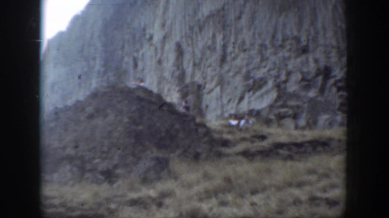 1937 California 8mm footage showing tiny hikers below a high rock face on a rugged mountain slope.