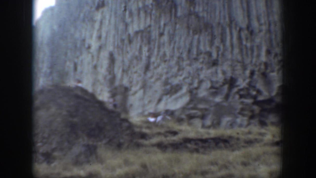 Rock wall and uneven grassy slope with small figures below.
