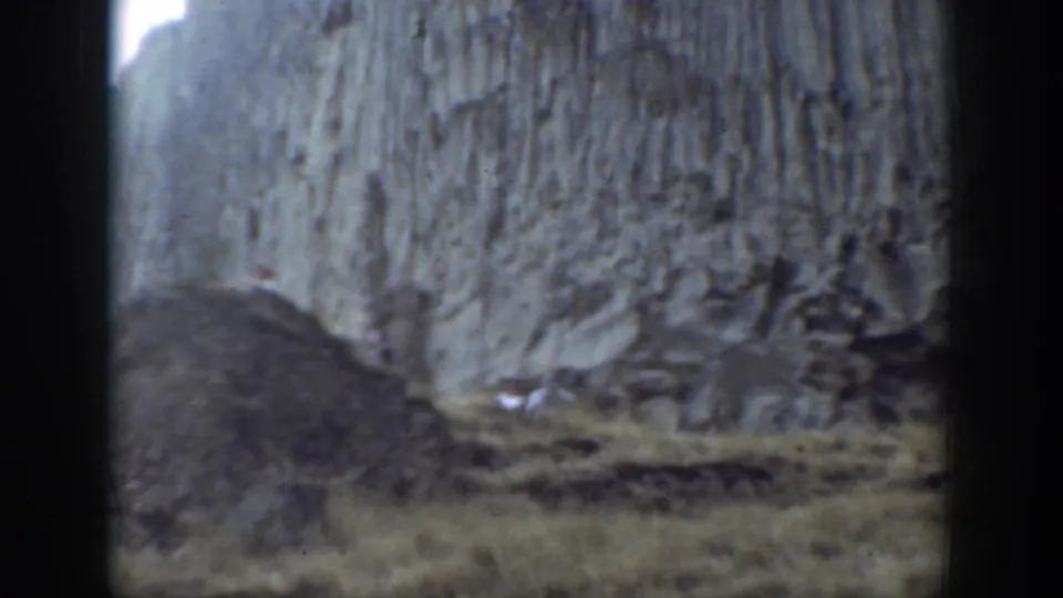 A distant view of hikers crossing rough terrain beneath a pale vertical rock wall in California home-movie footage.