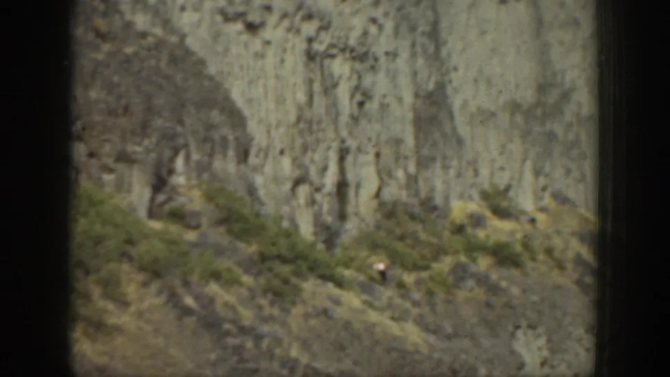 The camera looks toward a textured cliff and scrubby mountain slope, with a tiny person visible near the lower terrain.