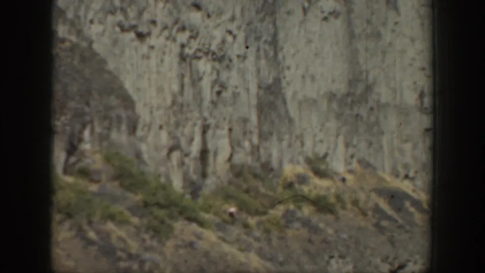 A faded 8mm view of a California mountain slope with a distant hiker beneath a sheer rock face.