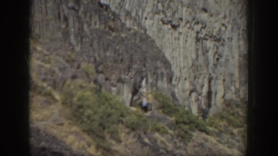 1937 California home-movie frame with a woman on a rugged hillside path against vertical rock formations and sparse brush.