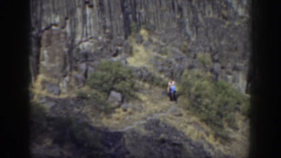 1937 California home-movie frame of a woman near the lower trail, surrounded by scrub, dry slope, and tall rock formations.