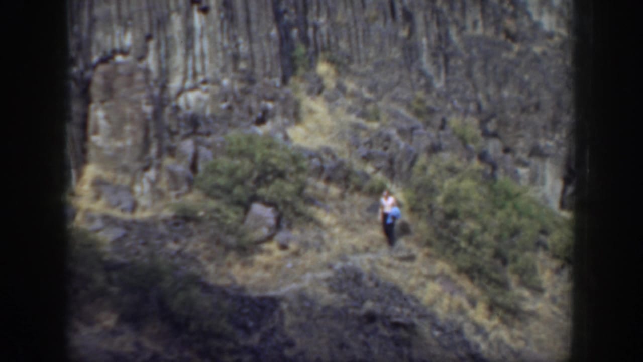 1937 California 8mm frame showing a lone walker on a dry trail beneath rugged dark rock and scattered shrubs.