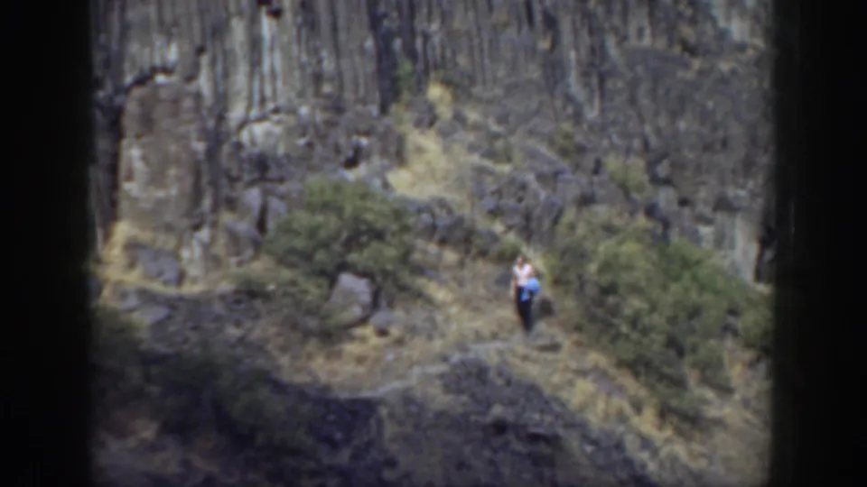 1937 California 8mm frame showing a lone walker on a dry trail beneath rugged dark rock and scattered shrubs.