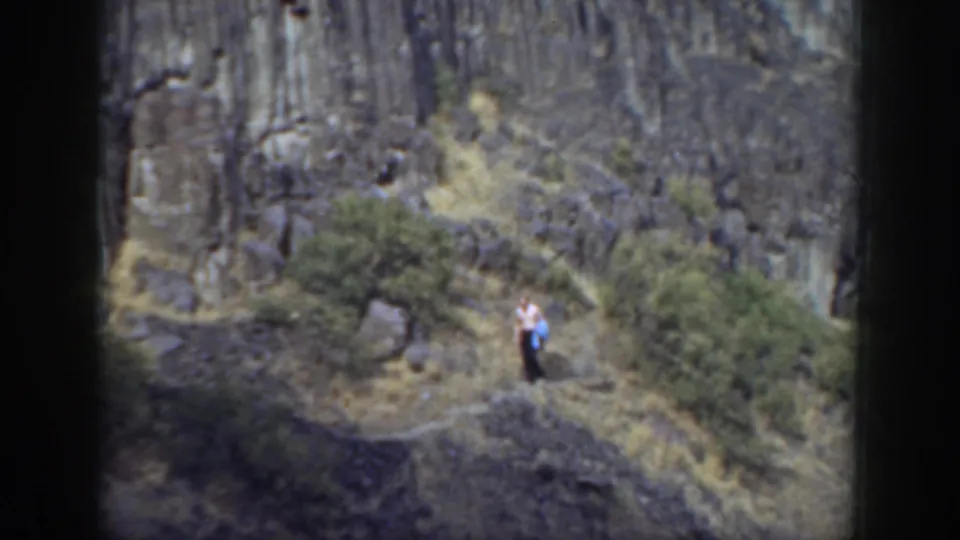 1937 California home-movie frame with a distant adult walking a sloped trail through dry grass and shrubs near dark rocks.