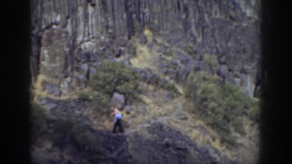 1937 California home-movie frame of a lone walker with a blue jacket crossing a trail below dark volcanic-looking rock.