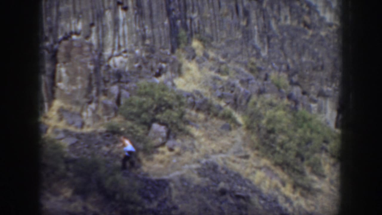 A distant hiker walks along a narrow California hillside trail beneath tall rock columns in 1937 home-movie footage.