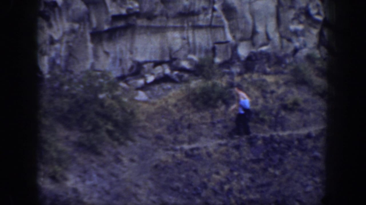 Hiker standing or moving on a brushy slope near dark rock formations.