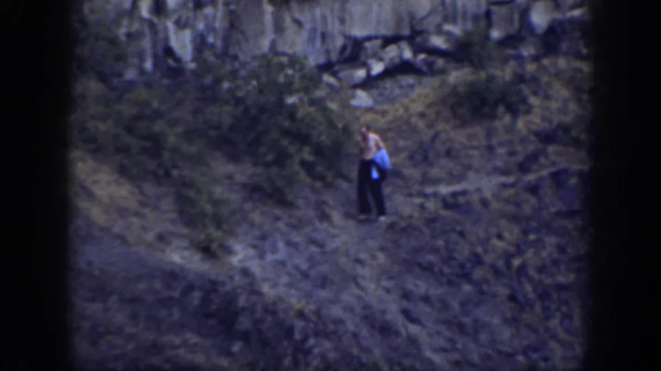 The hiker pauses or steps along a narrow hillside path with shrubs and rock ledges visible in the 1937 California home movie.