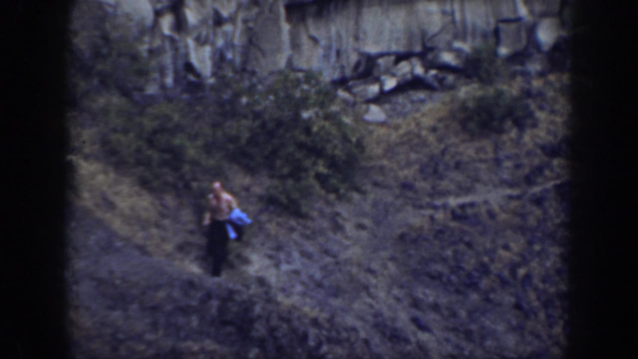 1937 California home-movie frame showing a distant shirtless man in dark pants near scrub below a rugged rock wall.
