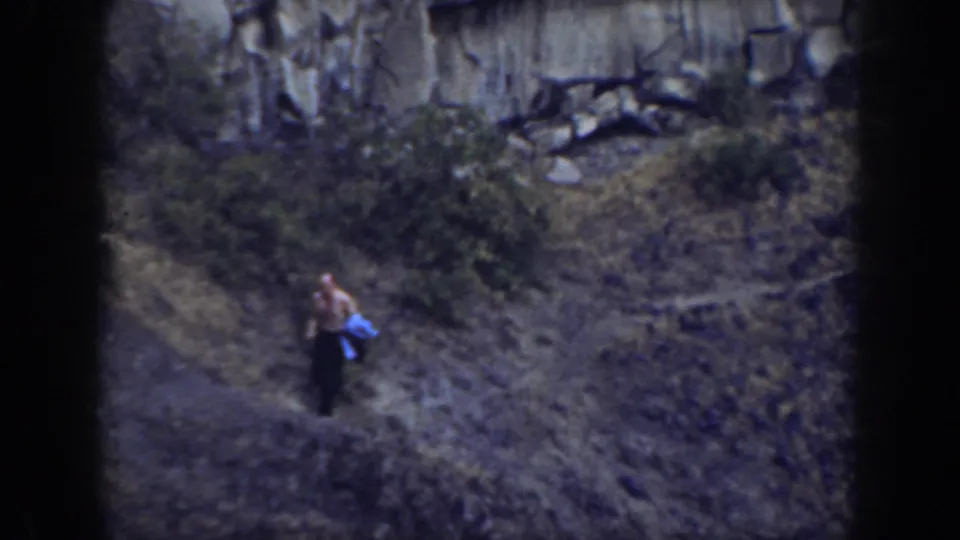 1937 California home-movie frame showing a distant shirtless man in dark pants near scrub below a rugged rock wall.