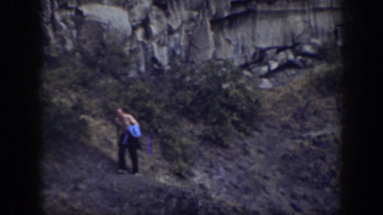 Man in dark pants stands on a narrow rocky path with scrub around him.