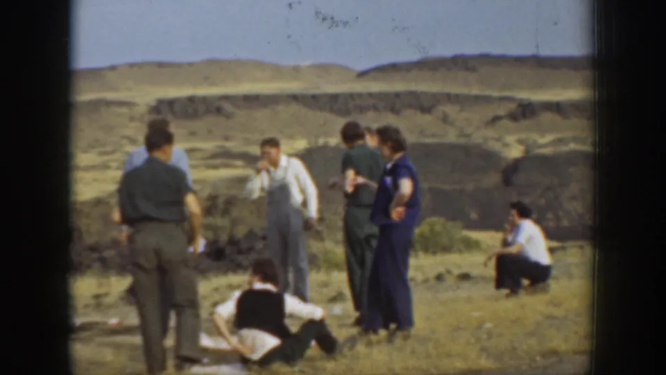 1937 California home-movie frame of adults gathered on rocky ground with dry hills in the distance.