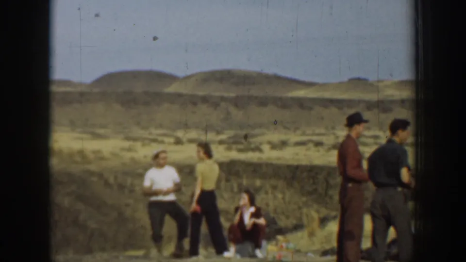 Adults in 1937 California stand and sit in a dry open landscape, with low hills and scrub visible behind them.