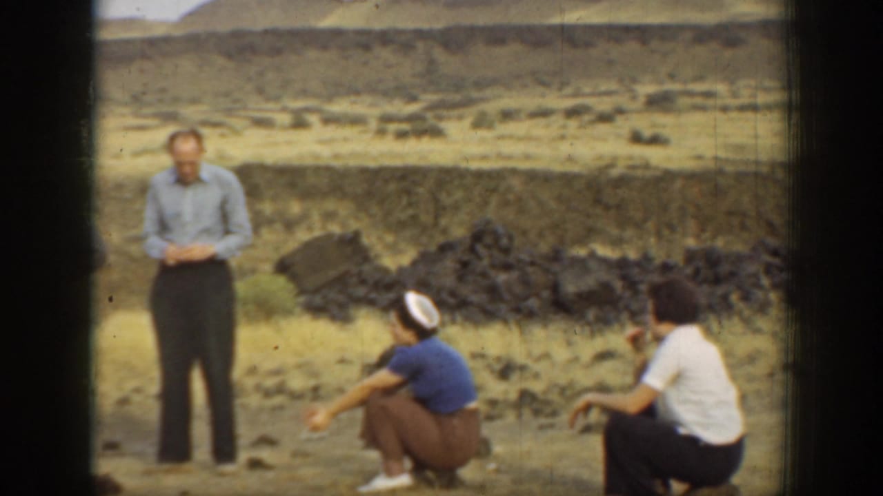 1937 California home-movie frame showing adults standing and sitting on dry rocky ground in a hilly landscape.