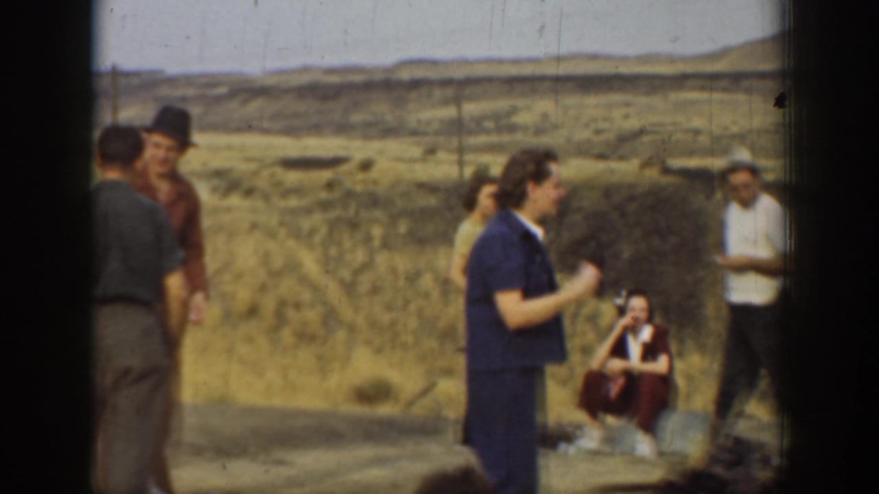 Adults in casual period clothing gather in a dry California hillside setting, with one person seated near the rocks.