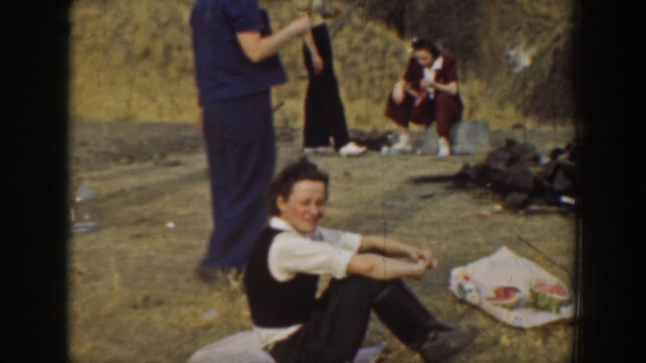A seated adult smiles toward the camera near picnic food on the ground, with other adults nearby in the dry hillside setting.