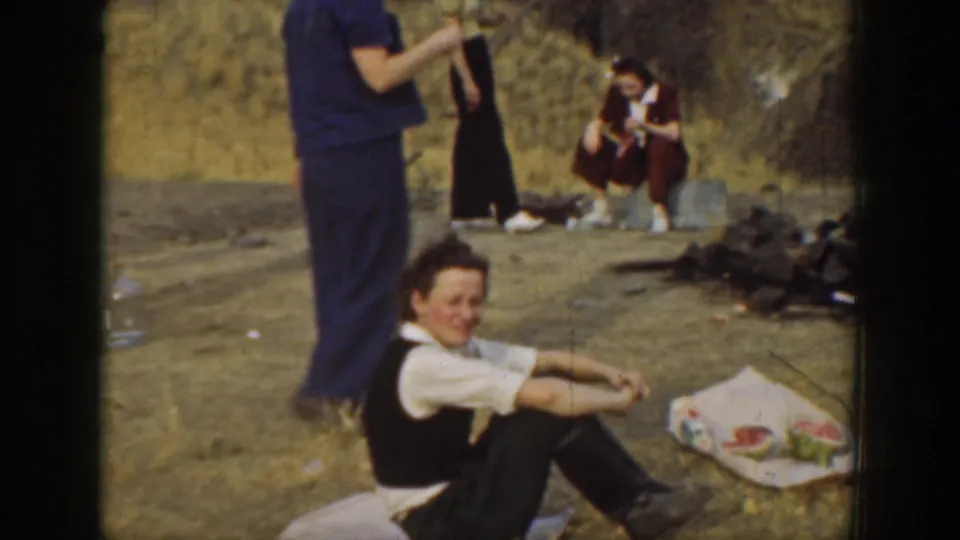 A seated adult smiles toward the camera near picnic food on the ground, with other adults nearby in the dry hillside setting.