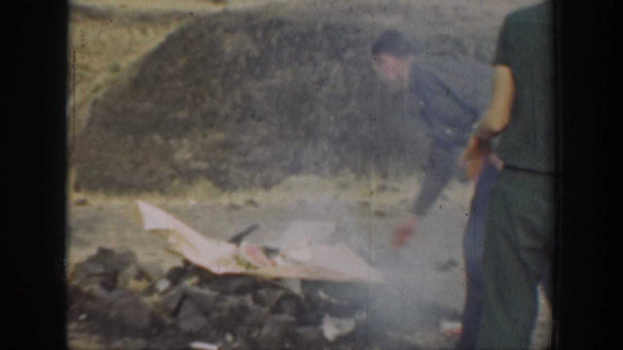 Smoke drifts from a small firepit or cooking area in the dry hillside setting while adults stand close by.