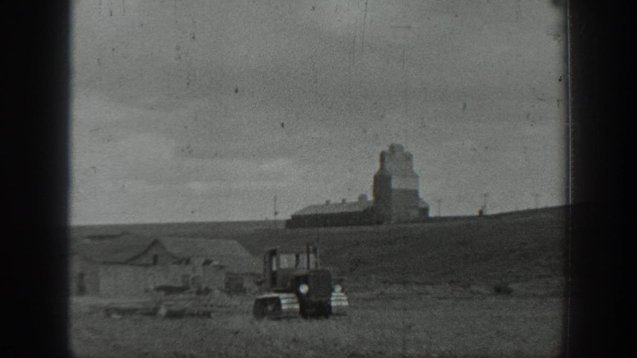 Tracked tractor in a field below distant farm buildings and a grain elevator.