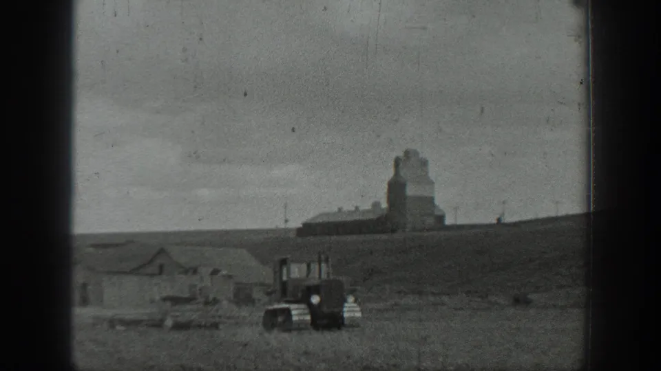 1937 California home-movie frame showing a tracked tractor in an open field with farm buildings and a grain elevator on a low hill.