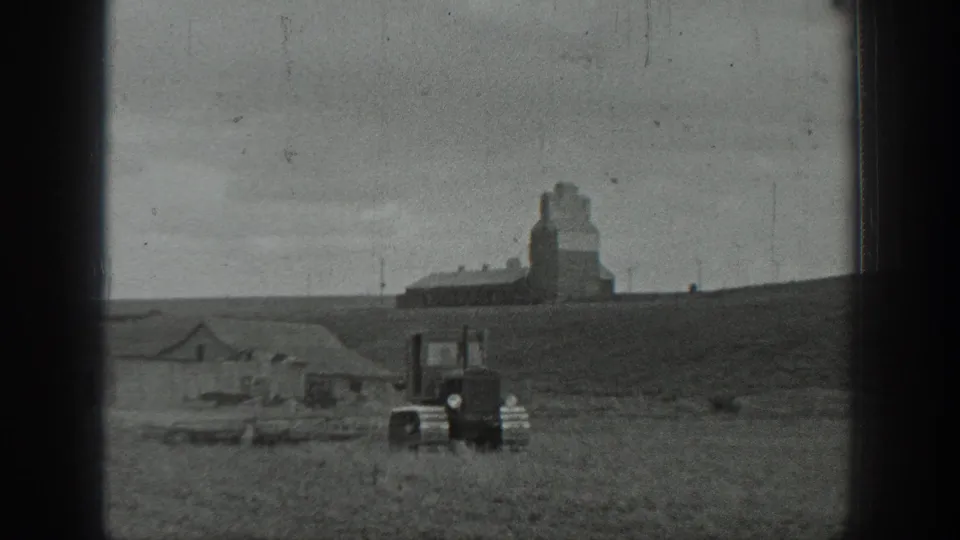 Tracked tractor seen at mid-distance in a rural California field, with a grain elevator and low buildings on the hillside beyond.