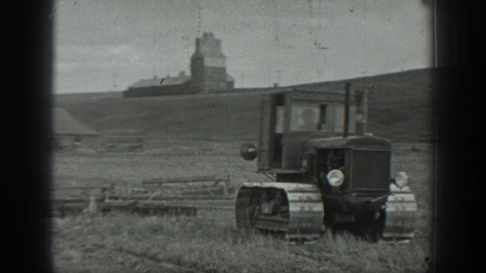 Closer 1937 rural California frame of a tracked tractor moving past fence rails, with farm buildings and a grain elevator in the distance.