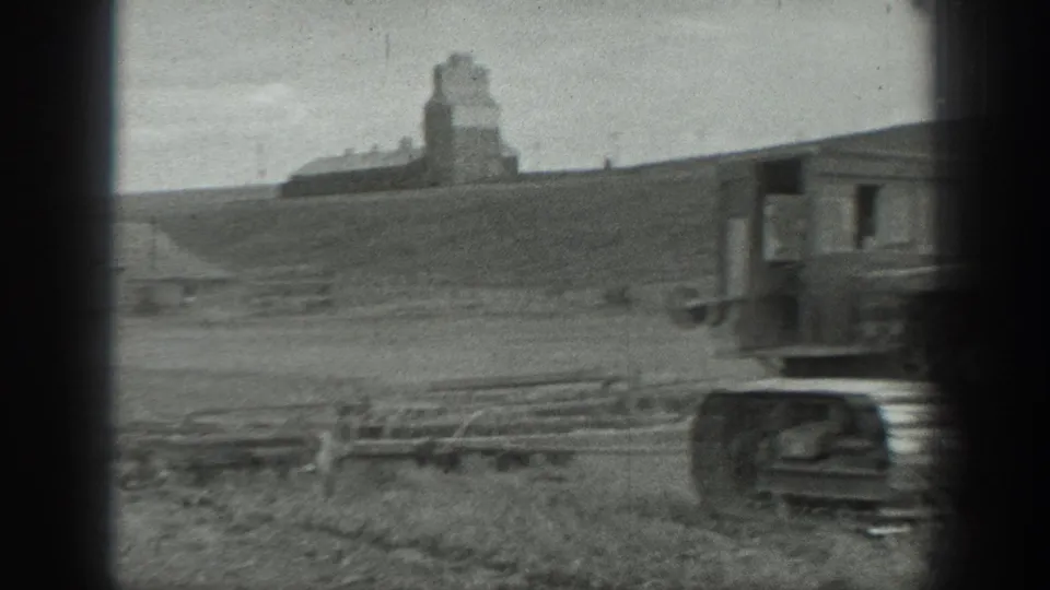 A tracked tractor moves partly out of frame in the field, leaving the distant grain elevator and rural hillside visible behind it.