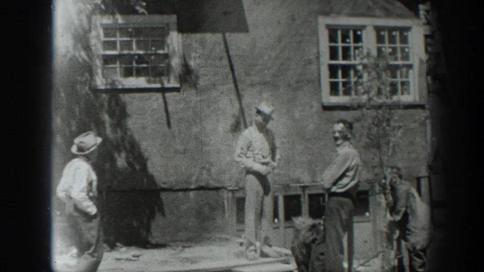 1937 California home-movie frame of men in hats and work clothes gathered beside a rustic building with windows and simple wooden rails.
