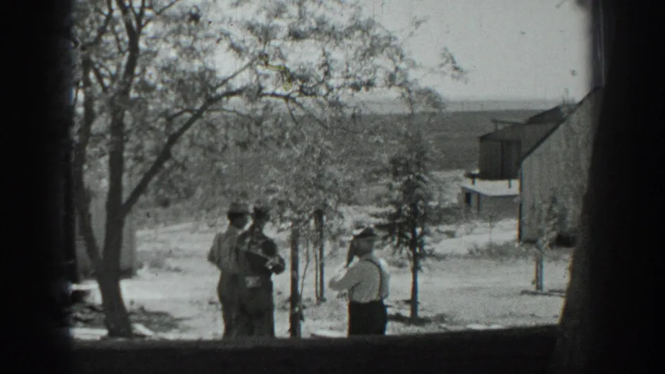 Three adults in a rural California setting in 1937 stand on a dirt road near trees, posts, a pale building, and distant hills.