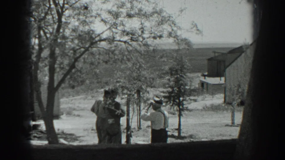 A 1937 California home-movie frame shows three adults conversing on a dirt road beside trees, with a building and hillside behind them.