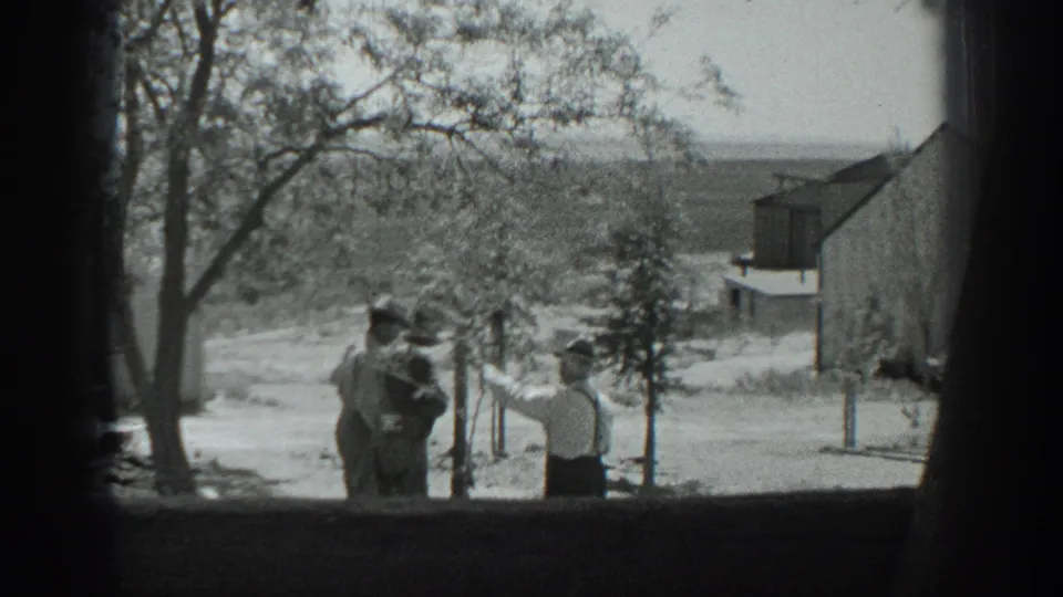 In the rural California yard, a man in a light shirt and suspenders gestures toward the trees as two men watch from the side.