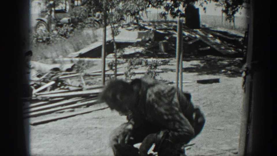 1937 California 8mm frame showing a person bending near lumber, debris, posts, and a low outdoor structure.