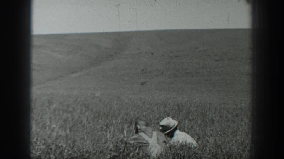 Black-and-white 1937 California home-movie frame with two distant workers in hats crouched in dense crops during a field pan.