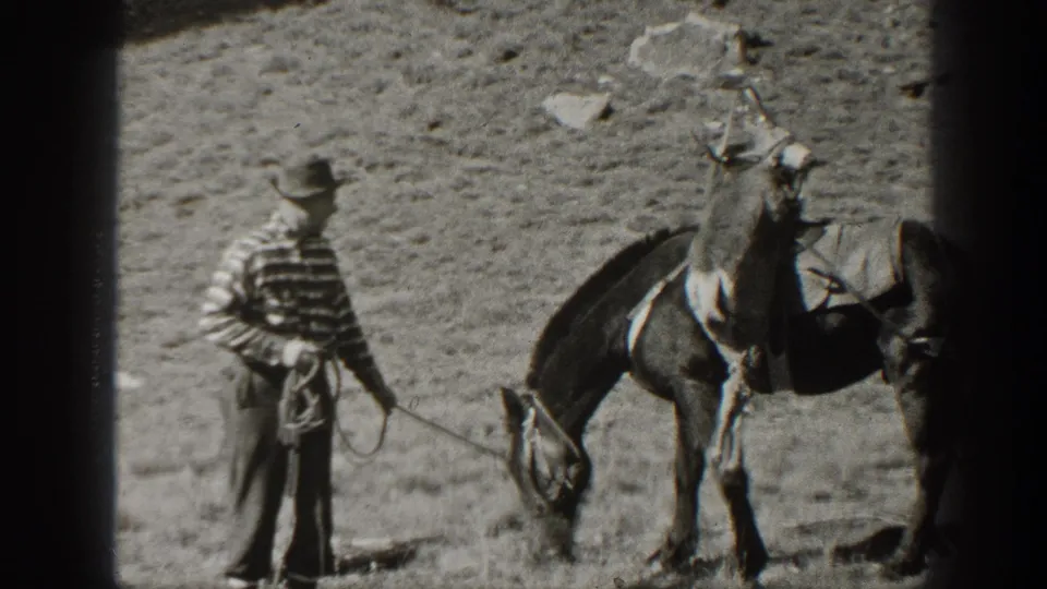 1938 California home-movie frame showing a man in a striped shirt leading a saddled pack mule across dry rocky terrain.