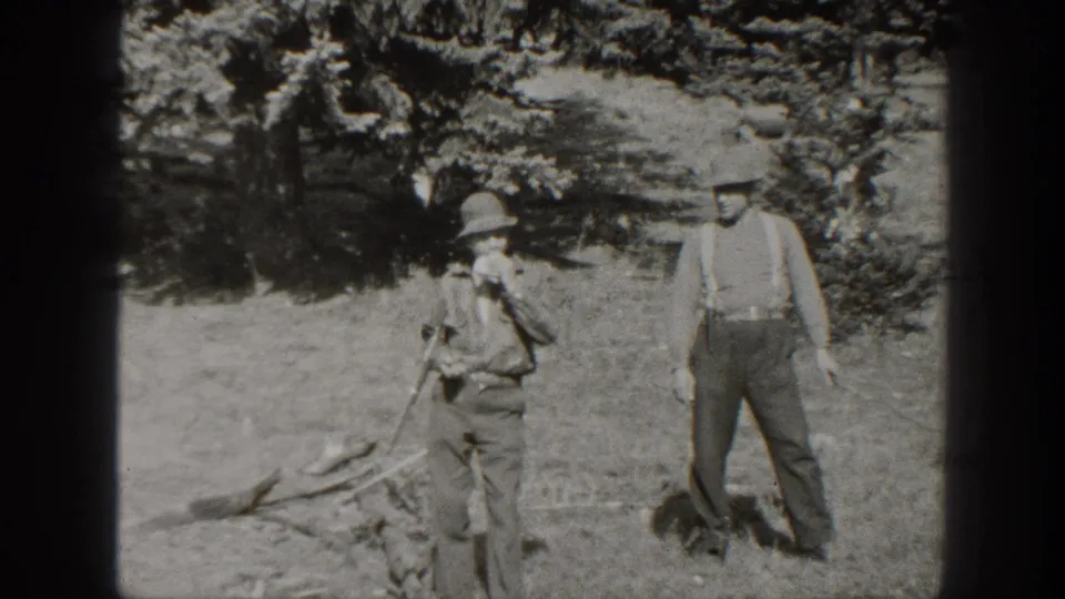 Later 1938 California frame showing two adult men in hats and suspenders standing in the same outdoor meadow area.
