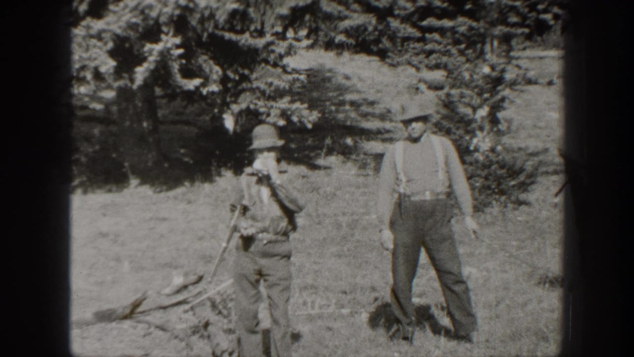 Two men stand in a wooded campsite, one holding a rifle and one wearing suspenders.