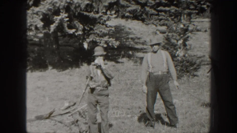 In 1938 California home-movie footage, two adult men stand on dry ground near trees, with one holding a rifle and the other in suspenders.
