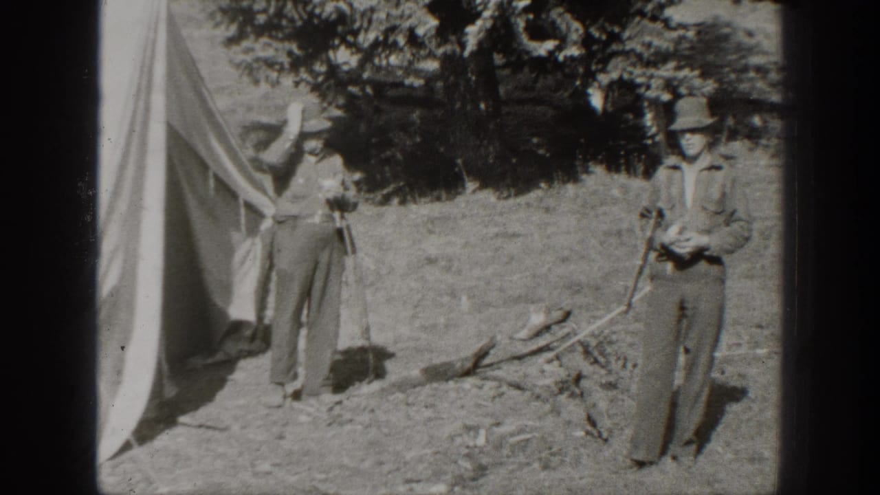 Two men stand near a canvas tent and scattered branches in a wooded clearing.