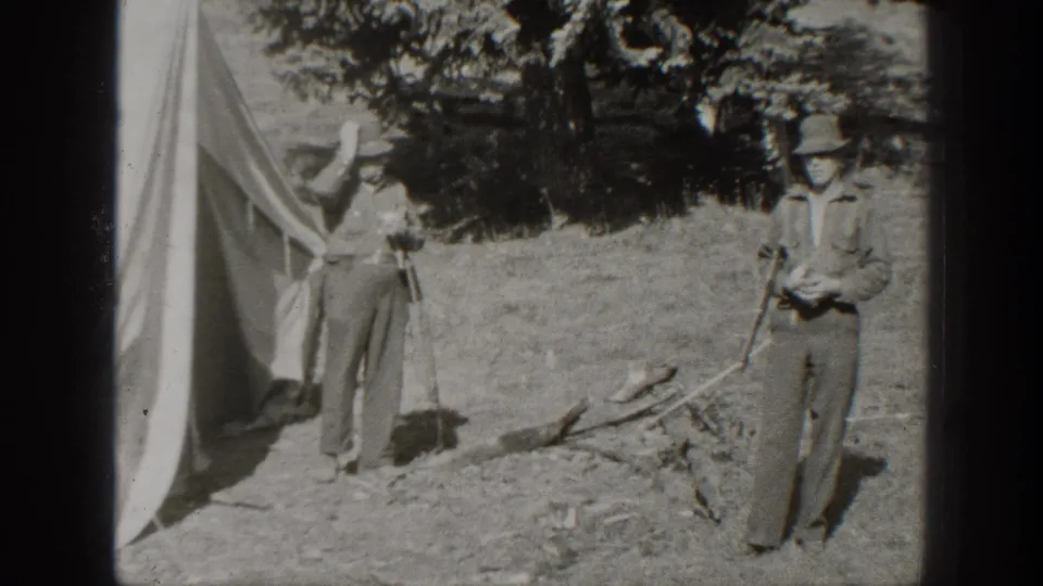Two adult men in hats stand beside a canvas tent in a California wooded campsite, with loose branches and dry ground nearby.