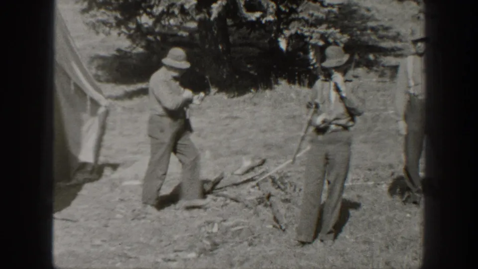 At the wooded campsite, one man stands near the tent while another holds a rifle, with dry brush and branches visible on the ground.