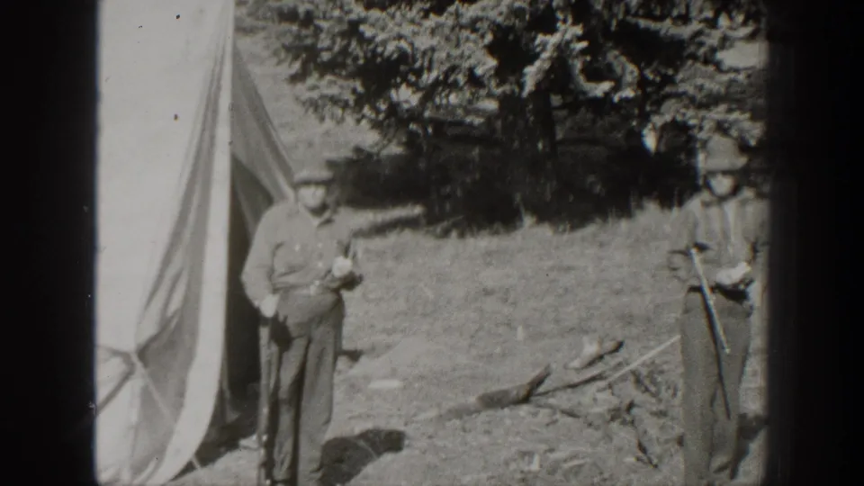 Two adult men stand near the edge of a canvas tent in 1938 California footage, with one man holding a rifle beside scattered camp wood.