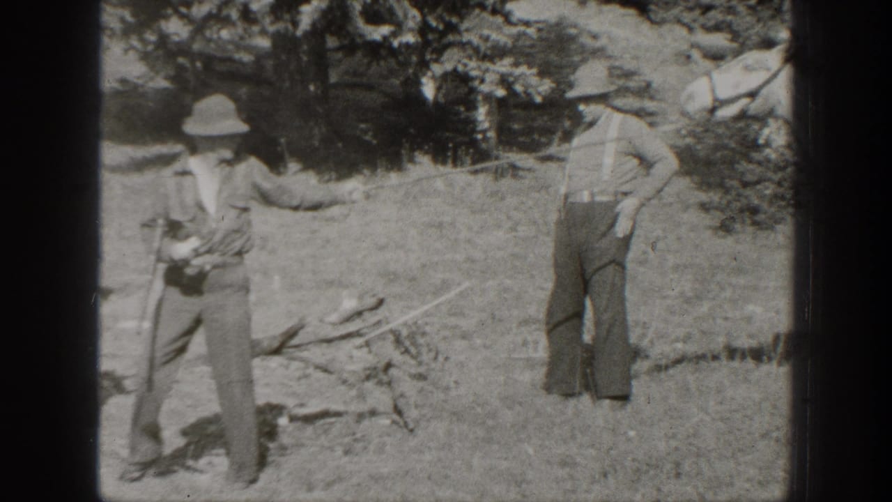 In 1938 California home-movie footage, two men in hats stand in a wooded camp clearing with grass and scattered supplies visible.