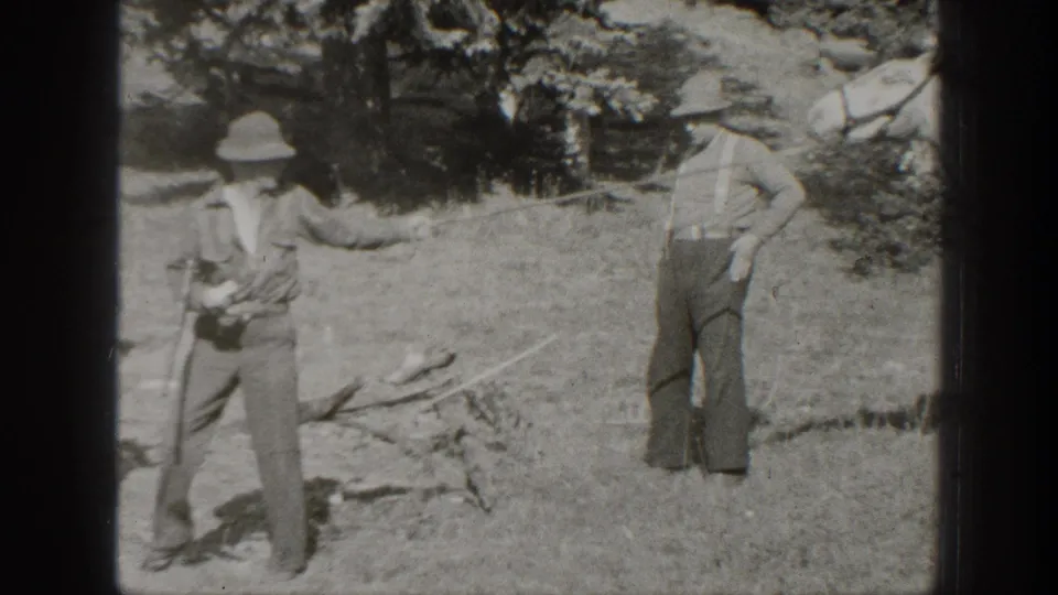 In 1938 California home-movie footage, two men in hats stand in a wooded camp clearing with grass and scattered supplies visible.