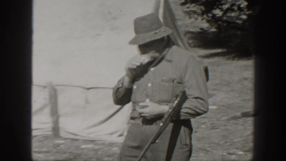 1938 California frame of a man in outdoor clothing beside a canvas tent, holding a rifle while eating.